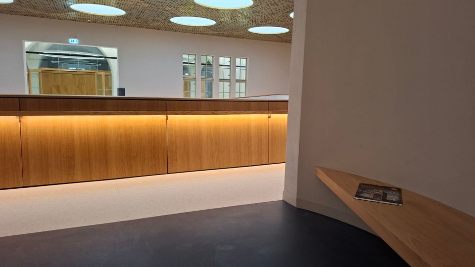 Moderner Innenraum mit langer, indirekt beleuchteter Holztheke vor einer weißen Wand mit großen Fensteröffnungen und Tür. An der Decke sind runde Lichtfelder in einer perforierten Deckenverkleidung, rechts befindet sich eine schmale Holzbank an der Wand.