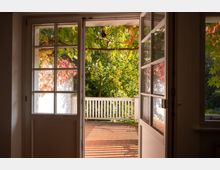 Blick aus einem Innenraum durch offene, verglaste Doppeltüren auf eine sonnige Holzterrasse mit weißem Geländer. Draußen sind dichter, grün‑ und rötlich gefärbter Laubbewuchs sowie Schattenmuster auf den Terrassendielen zu sehen.