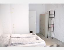 Schlichtes Hostelzimmer mit weiß gestrichenen Wänden, zwei nebeneinander stehenden Betten mit gemusterten grauen Bettdecken, einem Metallspind, einer Leiter und aufgerollten Handtüchern auf den Betten.