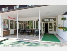 Eingangsbereich eines Hotelgebäudes mit überdachter Terrasse, auf der kleine Tische und Stühle stehen. Links befindet sich die Rezeption mit Glasfenstern und einer Tür, rechts die Eingangstür zum Hotel, beide von Grünpflanzen umrahmt.