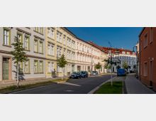 Straße mit sanierten Altbauten in Pastellfarben, teilweise mit grünen Fensterrahmen und roten Ziegeldächern, gesäumt von jungen Bäumen und Laternen. Am Straßenrand sind parkende Autos und Gehwege mit Grünstreifen zu sehen.