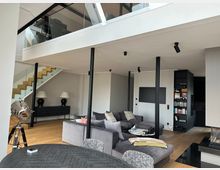Offener, moderner Wohnbereich mit hoher Decke und Galeriegeschoss mit Glasgeländer, verbunden durch eine Holztreppe. Im Raum stehen ein großes graues Ecksofa, ein runder Esstisch und schwarze Stützsäulen; rechts sind ein Einbau-Regal und eine schwarze Küchenzeile zu sehen.