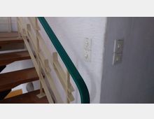 Ein Treppenaufgang mit Metallgeländer und Holzstufen, daneben eine weiß verputzte Wand mit zwei Schalterdoppelgruppen.