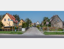 Wohnsiedlung mit Einfamilienhäusern in einer ruhigen Straße; links gelbe Häuser mit roten Dächern, rechts ein graues Haus mit Garten. Ein Straßenschild "30 Zone" und ein Schild mit Straßennamen ("Curlandstraße") markieren die Umgebung.