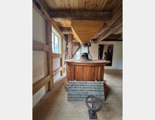 Innenraum einer historischen Mühle mit Holzbalken und einer hölzernen Getreidemühle. Im Vordergrund befindet sich eine handgeschriebene Tafel mit Hinweisen und ein eisernes Handrad, während natürliches Licht durch ein Fenster von der Seite einfällt.