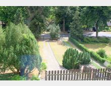 Gartenansicht mit mehreren Bäumen, Sträuchern und einem gepflasterten Weg, der zu einem Tor führt, das an eine ruhige Straße grenzt. Im Vordergrund ist ein Holzzaun sichtbar, im Hintergrund stehen hohe Bäume und Hecken.