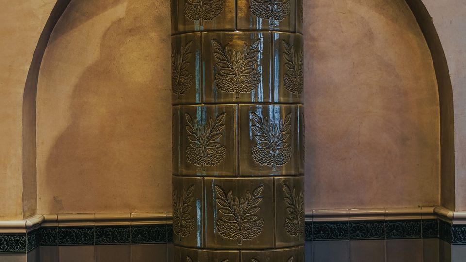 Historischer Innenraum mit einem hohen, zylindrischen Kachelofen in einer Wandnische, verziert mit reliefierten Blattmotiven und einer gewölbten Ofenhaube. Die Wand ist zweifarbig gehalten, unten mit Fliesen und oben mit glattem Putz.