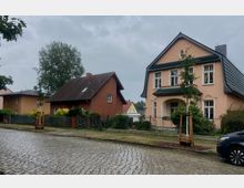 Zwei Einfamilienhäuser in einer ruhigen Wohnstraße mit Kopfsteinpflaster. Links ein rotes Backsteinhaus mit Satteldach, rechts ein beigefarbenes Haus mit grünem Walmdach und einer überdachten Eingangsterrasse; im Vordergrund frisch gepflanzte Jungbäume und Gehwege.