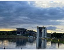 Wasserstraße mit einem Schiffshebewerk im Vordergrund, bestehend aus zwei hohen weißen Betontürmen mit gelben Beschriftungen. Links im Hintergrund eine Metallbrücke und eine Waldlandschaft, darüber ein bewölkter Himmel bei Sonnenuntergang.