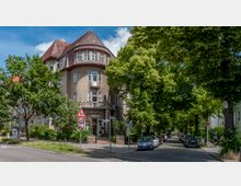 Straßenecke mit einer historischen Wohnhausfassade mit Erker und rotem Spitzdach, im Erdgeschoss ein Friseursalon. Rechts eine von Bäumen gesäumte Straße mit parkenden Autos und Fußgängern, links Verkehrszeichen und Grünflächen.