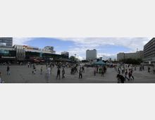 Weitläufiger Platz in einer urbanen Umgebung mit vielen Menschen, flankiert von modernen Gebäuden mit Geschäften und Restaurants, darunter ein Burger King und Saturn. In der Mitte des Platzes steht ein Pavillon mit einem grünen Dach, im Hintergrund sind weitere Bürogebäude und Bäume zu sehen.