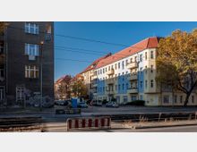 Straßenszene mit mehrstöckigen Wohngebäuden, eines davon in blauer und gelber Fassade mit roten Ziegeldächern. Im Vordergrund sind Straßenbahngleise, eine Absperrung und ein Baum mit Herbstlaub zu sehen.