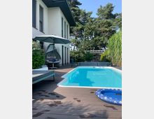 Modernes Wohnhaus mit hellem Putz und großen Fenstern, angrenzend an eine Terrasse mit Holzboden, einem rechteckigen Pool und Gartenmöbeln. Im Hintergrund befinden sich Bäume und eine kleine Überdachung, umgeben von dichter Vegetation.