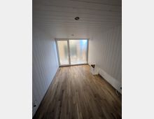 Schmaler, leerer Innenraum mit weiß vertäfelten Wänden und Decke sowie Holzfußboden. Am Ende befindet sich eine große, milchig verglaste Schiebetür bzw. Fensterfront, die Tageslicht in den Raum lässt.