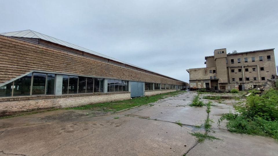 Ein verlassener Industriekomplex mit einer langen Halle aus Ziegelmauerwerk und großen Fenstern links sowie einem mehrstöckigen, verwitterten Gebäude rechts. Der Bereich dazwischen ist eine betonierte Freifläche mit vereinzeltem Grasbewuchs.