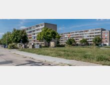 Wohngebiet mit mehrstöckigen Plattenbauten, Grünfläche im Vordergrund und Gehweg auf der linken Seite. Die Gebäude haben Balkone, teils mit Markisen oder Wäsche, und dazwischen stehen Bäume sowie eine kleine dekorative Wand mit einem Relief.