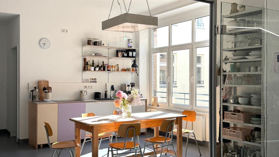 Heller Gemeinschaftsraum mit Küchenzeile und offenen Regalen, großem Holztisch mit Stühlen und einer hängenden Leuchte darüber. Große Fenster lassen viel Tageslicht herein; rechts steht ein verglaster Geschirrschrank, der Raum ist durch Glaswände abgetrennt.