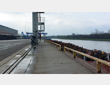 Ein Hafenbereich mit Betonpier am Fluss, rechts beladen mit Schrott auf einem Schiff. Links ein Lagergebäude und Kräne, im Hintergrund Industrieanlagen und Bäume entlang des Ufers.