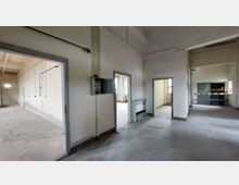 Ein leerer Raum in einem industriellen oder verfallenen Gebäude mit weiß gestrichenen Wänden, sichtbaren Kabeln und offenen Türen, die in angrenzende Räume führen. Im Vordergrund ist ein alter Sicherungskasten zu sehen, und einige Bereiche wirken durch Absperrband abgetrennt.