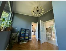 Wohnzimmer mit grauen Wänden, einem Holzparkettboden und einer großen Fensterfront links mit Pflanzen auf der Fensterbank. Im Raum steht ein Regal mit Brettspielen, und zwei offene weiße Doppeltüren führen zu einem hellen Bad und einem weiteren Raum.