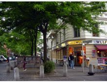 Straßenszene mit einem Eckgebäude, das eine Bäckerei und Konditorei („Wulf“) beherbergt. Vor dem beigen Gebäude stehen Bäume, mehrere Personen bewegen sich entlang des Bürgersteigs, und im Vordergrund sind Betonpoller sowie ein Stromkasten mit Graffiti sichtbar.