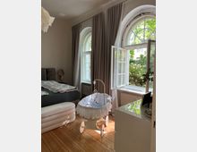 Schlafzimmer mit Holzboden und zwei großen, halbmondförmigen Fenstern, die von grauen Vorhängen umrahmt sind. Im Raum stehen ein Bett, ein weißer Stubenwagen mit Rüschen und ein weißes Sideboard; durch die geöffneten Fenster fällt Tageslicht auf das Innere.