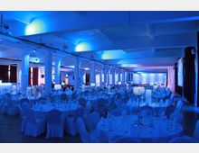 Großer Veranstaltungssaal mit runden, weiß gedeckten Tischen, geschmückt mit Kristall-Leuchtern als Mittelstücke. Die blaue Beleuchtung dominiert den Raum, der von Säulen und hohen Decken geprägt ist.