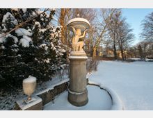 Schneebedeckte Parklandschaft mit einer steinernen Statue, die eine Schale trägt, auf einem hohen Sockel. Im Hintergrund winterliche Bäume, ein offenes Schneegebiet und ein Gebäude im warmen Licht der tief stehenden Sonne.