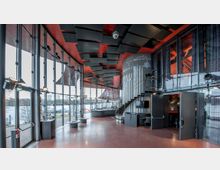 Modernes Foyer eines Veranstaltungsgebäudes mit großen Glaswänden, die den Blick auf einen Fluss und eine Außenlandschaft ermöglichen. Der Raum verfügt über rote Böden, eine geschwungene Treppe, Akustikelemente an der Decke und eine minimalistische Ausstattung mit Tischen und Beleuchtung.