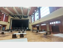 Ein Veranstaltungssaal mit einer Bühne im Hintergrund, mehreren weißen Tischen und Stühlen auf einem Holzboden. Große Fenster und rote Vorhänge lassen viel Tageslicht in den Raum, an den Wänden hängen Scheinwerfer.