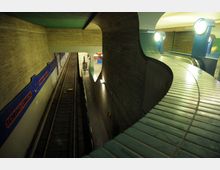 U-Bahn-Station mit Gleisen in einem Tunnel aus Betonwänden, beleuchtet durch moderne Lampen. Im Vordergrund sind geschwungene, mit grünen Fliesen verkleidete Geländer und eine Rolltreppe sichtbar.