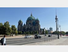 Straßenansicht mit dem Berliner Dom im Zentrum, einem historischen Gebäude mit großen grünen Kuppeln. Rechts im Hintergrund ist der Fernsehturm sichtbar, flankiert von modernen und historischen Gebäuden sowie Bäumen entlang der Straße.