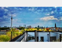 Blick auf die Skyline von Berlin mit dem Fernsehturm im Vordergrund, umgeben von weiteren markanten Gebäuden wie dem Berliner Dom, aufgenommen von einer begrünten Dachterrasse mit Metallgeländer. Der Himmel zeigt dichte Wolken und vereinzelte blaue Stellen.