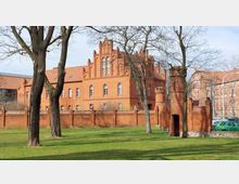 Backsteinbau im neugotischen Stil mit spitzbogigen Fenstern und Zinnen, umgeben von einer Backsteinmauer und einem begrünten Parkbereich mit Bäumen. Im Vordergrund steht ein kleiner, turmförmiger Anbau der Mauer.