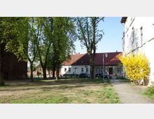 Gartenfläche mit großen Bäumen und gepflastertem Weg, gesäumt von einem gelben Blütenstrauch rechts. Im Hintergrund befinden sich Häuser mit roten Ziegeldächern und parkende Autos vor der Fassade.
