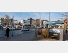 Eingang zur U-Bahn-Station "Rudow" der Linie U7 in Berlin mit einem blauen Stationsschild, umgeben von einer belebten Straße, mehreren Autos, Fußgängern und modernen Wohn- sowie Geschäftshäusern im Hintergrund. Links sind Straßenlaternen, Wegweiser und Fahrradständer zu erkennen.