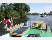 Ein Fluss mit einem kleinen Boot im Vordergrund, dessen Deck mit bunten Kissen ausgelegt ist; im Hintergrund Ufer mit Bäumen sowie industrielle Bauwerke mit einem hohen Schornstein.