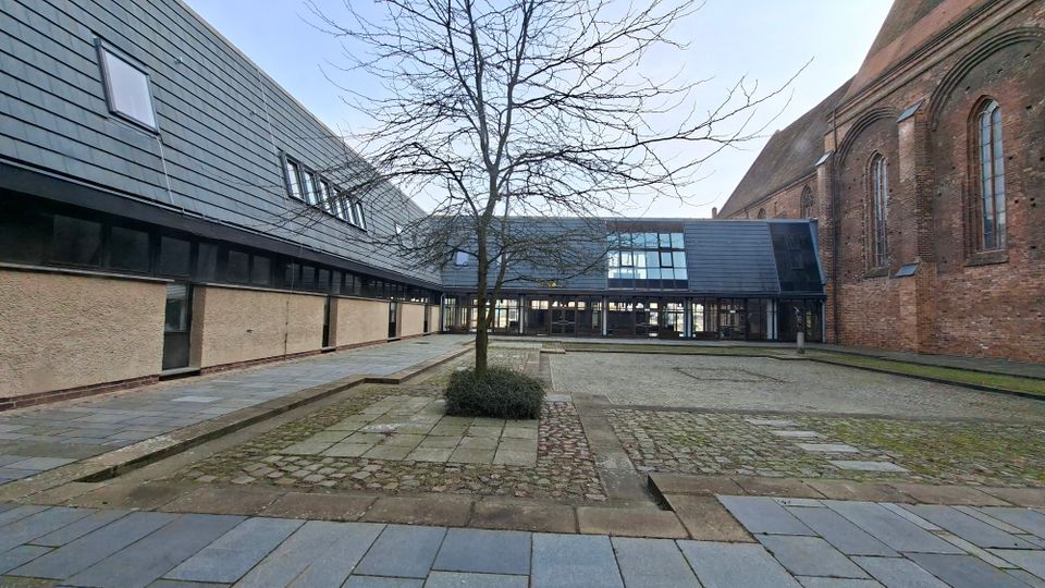 Innenhof zwischen einem modernen, dunkel verkleideten Gebäude und einer großen Backsteinkirche mit hohen Rundbogenfenstern. Der gepflasterte Platz hat breite Steinwege, niedrige Stufen und einen zentralen, blattlosen Baum vor einer Glasfassade am Hofende.