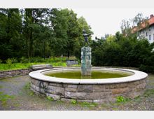 Ein runder Brunnen aus Stein mit grünlichem Wasser, in dessen Mitte eine Statue eines Kindes auf einem steinernen Sockel steht. Der Brunnen befindet sich in einer parkähnlichen Umgebung mit Bäumen, Sträuchern und einer angrenzenden Sitzbank.