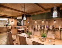 Ein modernes Restaurant im rustikalen Stil mit Tischen, Stühlen und Wänden aus hellem Holz. Im Vordergrund stehen Tische mit Kräutertöpfen, und im Hintergrund ist eine offene Küche mit einem Koch zu sehen.