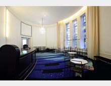 Innenraum eines modern gestalteten Cafés oder Warteraums mit abgerundeter Wand, hohen Fenstern mit farbigem Glas, hellen Vorhängen und einer dunklen Theke. Der Boden ist mit blau gemustertem Teppich bedeckt, und es stehen weiße Tische sowie Stühle mit unterschiedlichem Design.