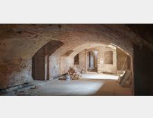 Ein Kellerraum mit Gewölbedecke aus Ziegelsteinen, roh belassene Backsteinwände und unbefestigter Boden mit Baumaterialien, darunter ein Haufen Ziegelsteine und ein Farbeimer. Licht fällt durch eine Öffnung und ein kleiner Durchgang ist im Hintergrund erkennbar.