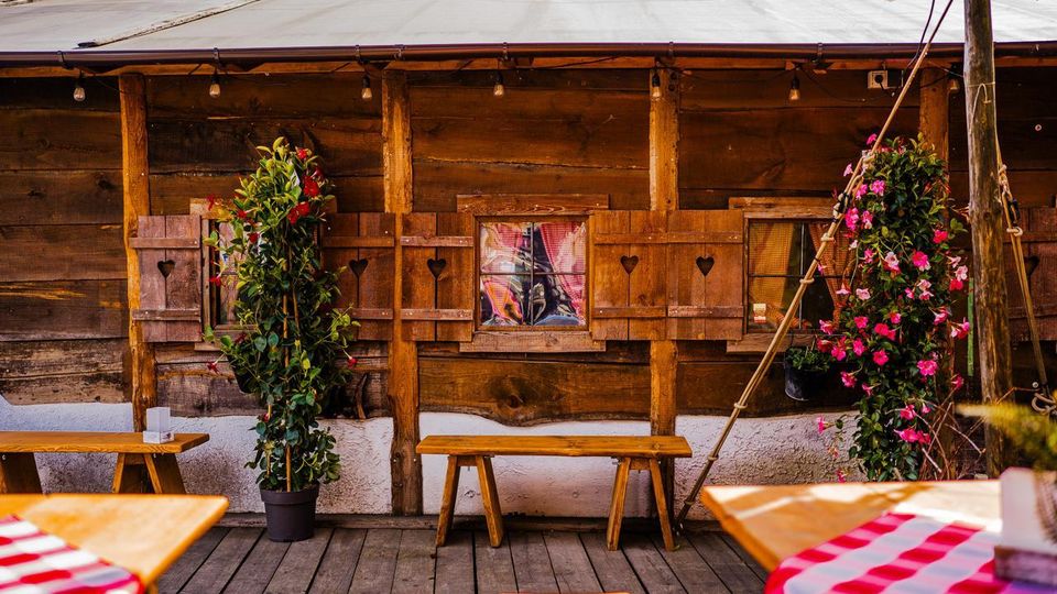 Rustikale Holzfassade einer Hütte mit kleinen Fenstern und Fensterläden mit herzförmigen Ausschnitten sowie einer Lichterkette unter dem Dach. Davor stehen Holzbänke und Tische auf einer Terrasse, flankiert von großen Topfpflanzen und blühenden Kletterpflanzen.