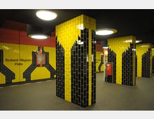 U-Bahn-Station mit gelb-schwarzen Wand- und Säulenfliesen, die abstrakte Muster bilden, beleuchtet durch runde Deckenlampen; an einer Wand ist der Schriftzug "Richard-Wagner-Platz" zu sehen.