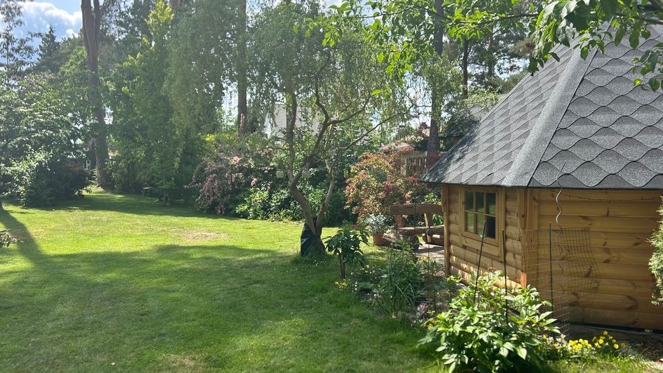 Großer, gepflegter Garten mit weitläufiger Rasenfläche, umgeben von hohen Bäumen, Sträuchern und blühenden Beeten. Rechts steht ein kleines Holz-Gartenhaus mit schindelgedecktem, steilem Dach und Fenster, daneben eine Terrasse mit Pflanzen.
