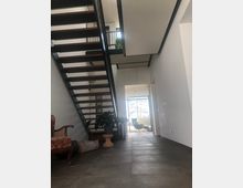 Modernes, minimalistisches Interieur mit einer offenen Metall- und Holztreppe, großformatigen grauen Bodenfliesen und einer Sitzgruppe nahe einer großen Glasfront mit Blick auf Bäume im Hintergrund. links ein gepolsterter Stuhl und Zimmerpflanzen.