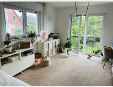 Helles Wohnzimmer mit Teppichboden, großen Fenstern und einer Glastür zum Balkon mit Blick auf einen Garten. Die Einrichtung umfasst weiße Regalmodule, Zimmerpflanzen und Dekor wie eine rosafarbene Lampe sowie ein Sitzpouf.