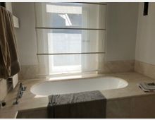 Helles Badezimmer mit einer eingelassenen Badewanne aus beigem Marmor, einem darüber liegenden Fenster mit halbtransparentem Rollo, einem aufgerollten Handtuch und einer Zeitung auf der Ablage.