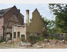 Teilweise eingestürztes Gebäude aus rotem und gelbem Backstein mit sichtbaren Schäden und verfallenen Fassaden. Im Vordergrund befinden sich Trümmer und überwachsene Flächen, im Hintergrund Bäume.
