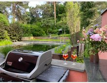 Terrasse in einem begrünten Garten mit modernem Naturpool, Holzterrassen und gestuften Beeten vor dichtem Baumbestand. Im Vordergrund stehen ein Gasgrill, zwei Getränkegläser und eine Vase mit Blumen neben einer Gartenmauer.